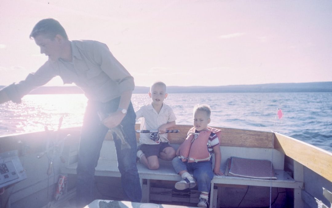 man holding gray fish standing beside two boys sitting on boat seats