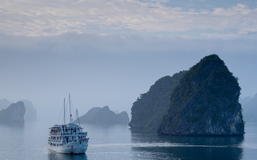 white ship on body of water near the rock formation covered with grass and tree