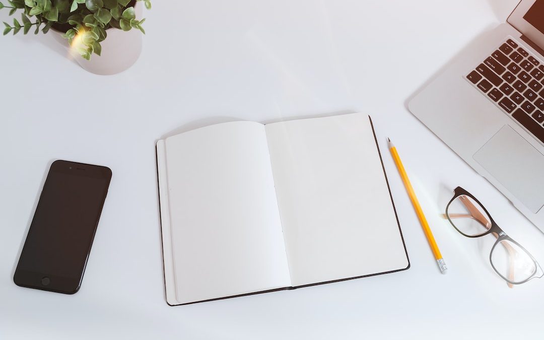 An open empty notebook on a white desk next to an iPhone and a MacBook