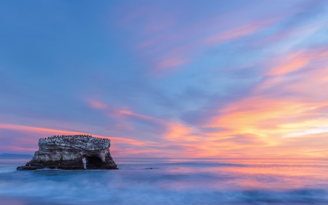 a large rock sticking out of the ocean under a colorful sky