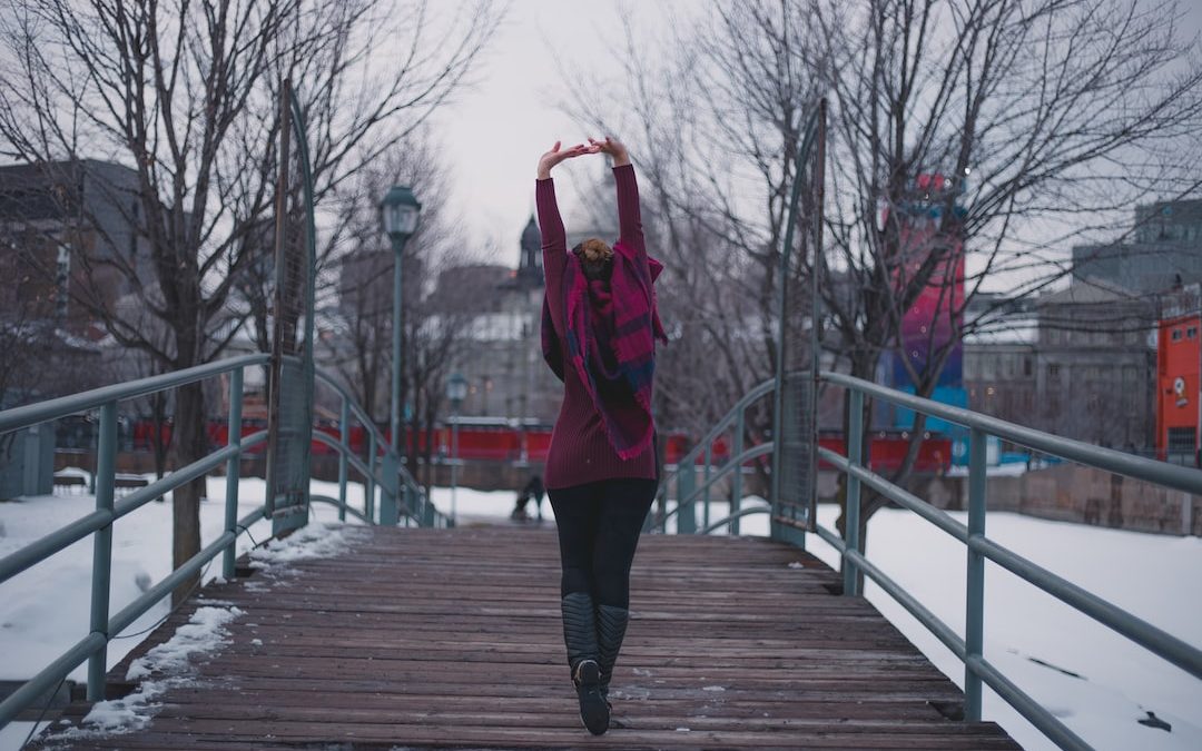 person wearing red jacket raising both hands while walking on brown wooden bridge during daytime