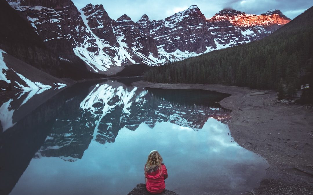 woman sitting on rock in front calm body of water and mountains view