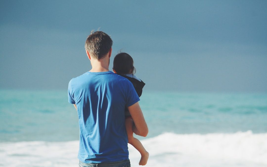 man wears blue crew-neck t-shirt holding toddler wears black hooded jacket near ocean under blue sky at daytime